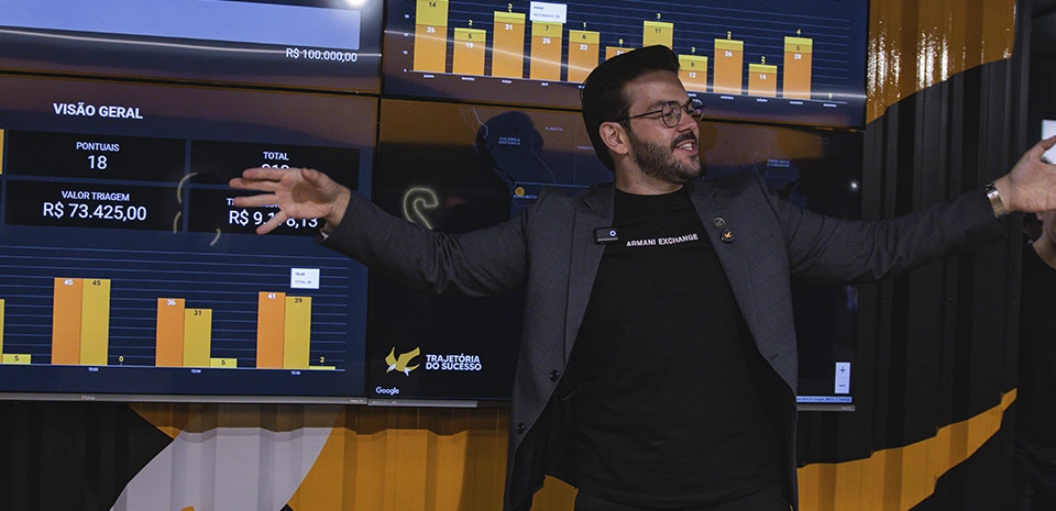 Renan Nicolini de braços abertos diante de painéis com indicadores de faturamento, em evento do Grupo TS