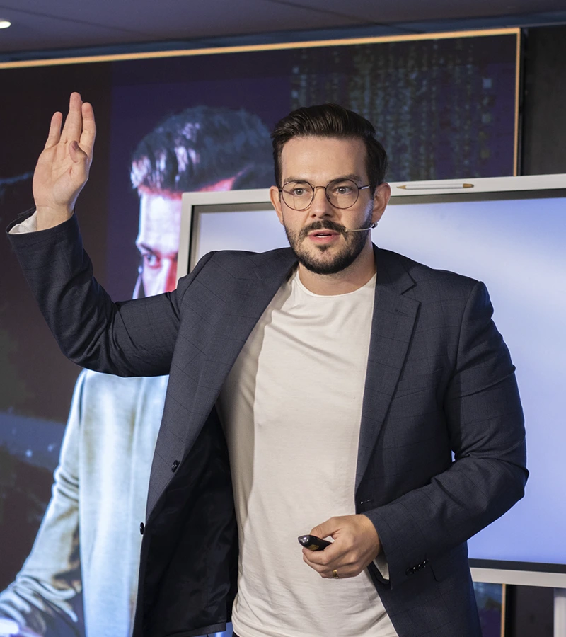 Renan Nicolini de microfone headset apresentando diante de um telão, com a mão erguida